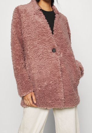Femme portant un manteau en fausse fourrure texturée rose poussière surdimensionné avec un bouton noir, un haut noir et un pantalon crème sur un fond blanc.