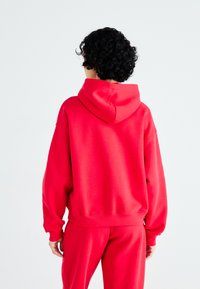 Hanorac roșu dintr-un material moale, cu o croială lejeră, având un guler cu șnur și manșete cu model în dungi, asociat cu pantaloni roșii asortați.