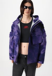 Purple tie-dye jacket with multiple front pockets, grey lining, and a hood. Worn over a black ribbed crop top with a red logo.