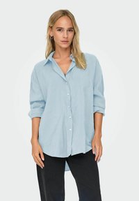 Camicia azzurra chiara a maniche lunghe con polsini arrotolabili, frontale con bottoni e una vestibilità rilassata. Realizzata in un tessuto morbido e testurizzato.