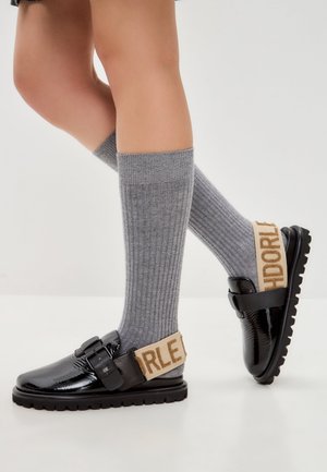 Musta lakkapinta loafers paksulla pohjalla, säädettävä solkitieto ja beige nauha kohokuvioiduilla kirjaimilla. Yhdistetty harmaisiin ribattuihin polvisukkiin.