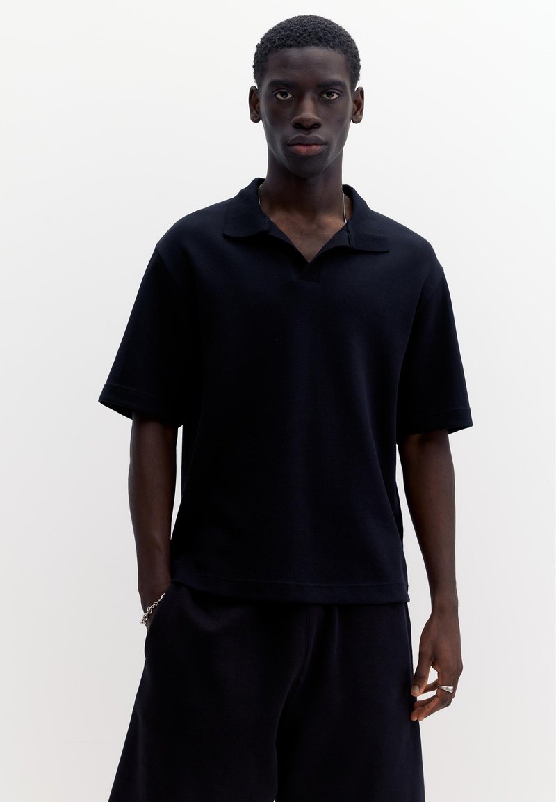 Jeune homme à la peau foncée portant un polo noir ample à manches courtes et un pantalon noir, debout devant un fond blanc uni.