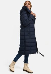 Navy puffer coat with a hood, featuring quilted design, zip closures, and side pockets. Worn with blue jeans and cream-colored boots.
