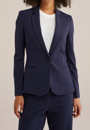Femme portant un blazer bleu marine ajusté avec un seul bouton sur un top blanc perforé, associé à un pantalon bleu marine assorti.