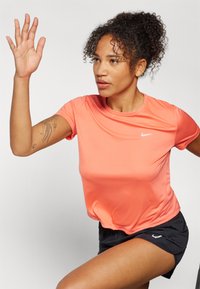 Idrettsantrekk bestående av en tettsittende koralloransje Nike T-skjorte med korte ermer, sammen med svarte shorts som har en jevn tekstur og logo.