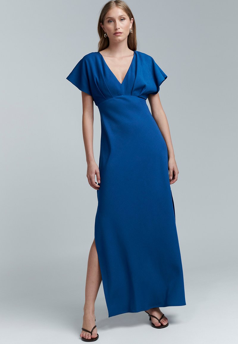 Dlhé priliehavé šaty z tmavomodrej látky s výstrihom do V a krátkymi rozviatymi rukávmi. Majú vysoký rozparok na jednej strane.