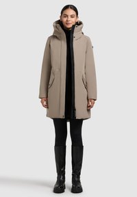 Woman wearing a taupe hooded winter coat, black leggings, and knee-high black boots, standing against a plain light gray background.