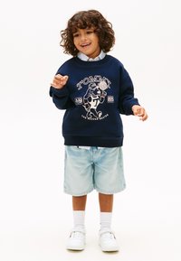 Ένα χαμογελαστό παιδί με κατσαρά μαλλιά φοράει σκούρο μπλε φούτερ με στάμπα σκύλου και μπάσκετ, ανοιχτόχρωμο τζιν σορτς, λευκές κάλτσες και αθλητικά παπούτσια.