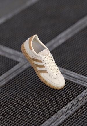 Biało-beżowy sneaker Adidas Spezial ze sznurowadłami zawieszony nad metalową kratą z przecinającymi się liniami.