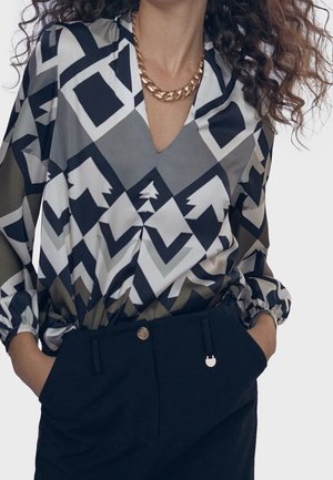 Mujer con blusa de manga larga de patrones en negro, blanco y gris con formas de chevrones y árboles, collar de cadena dorada y pantalones oscuros, con las manos en los bolsillos.