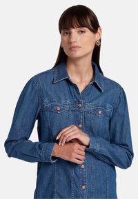 Denim overhemd in donkerblauw met knoopsluiting aan de voorkant, borstzakken en een getailleerde kraag. Heeft lange mouwen met knoopmanchetten.