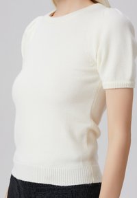 Haut en tricot blanc à manches courtes avec un col rond et un ourlet en côtes. Présente des manches bouffantes et une texture lisse et douce.