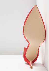 Chaussure à talon haut rouge positionnée debout avec un bout pointu et une semelle texturée visible sur fond blanc.