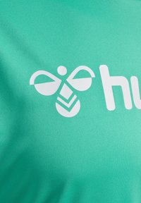 Turkusowa tkanina z białym logo o symetrycznym wzorze, zawierającym abstrakcyjne kształty przypominające pszczoły oraz wzór w zygzak.