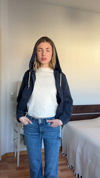 Νεαρή γυναίκα με κλειστά μάτια, φορώντας μπλε σκούρο μπουφάν με κουκούλα, λευκό πουκάμισο και μπλε τζιν, στέκεται μέσα σε εσωτερικούς χώρους κοντά σε κρεβάτι και κομοδίνο.