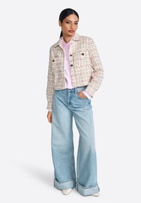 Giacca in tweed bianca con fili multicolori, dotata di due tasche frontali e bottoni dorati, abbinata a una blusa rosa chiaro e jeans a gamba larga.