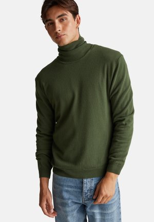 Suéter verde de cuello alto en material suave, con mangas largas, puños acanalados y un cuello ajustado, combinado con jeans de mezclilla azul claro.