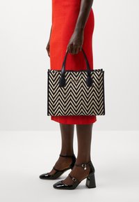 Vævet tote bag med et chevron-mønster i sort og beige, som har sorte læderhåndtag og detaljer med guld logo. Egnet til daglig brug.