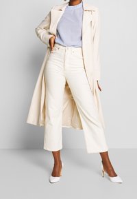 Beige lång kappa, ljusblå ribbad topp, krämfärgade vida jeans och vita högklackade skor. Kappan har slagkrage och sidfickor.