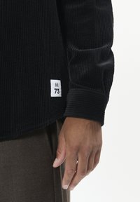 Sort corduroy skjorte med lange ærmer og ribbet tekstur, med et hvidt mærke med "M" og "73" i kanten, båret med brune bukser.