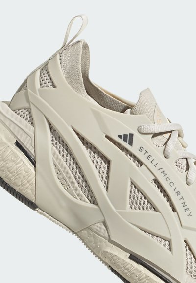 Zapatilla deportiva beige con parte superior de malla y refuerzos estructurados, suela texturizada y un sutil logotipo en el costado y la lengüeta.