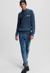 Pull bleu côtelé à col haut, ornée d'une broderie de logo blanche, associé à un jeans bleu délavé et des bottes noires massives.