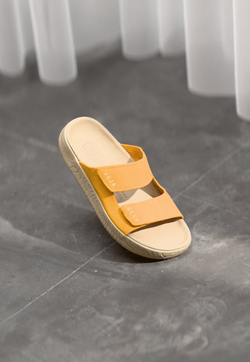 Keltaiset liukusandaalit, joissa on kaksi säädettävää hihnaa, kuvioitu pohja ja sileä pohja. Hihnassa on brändäys.