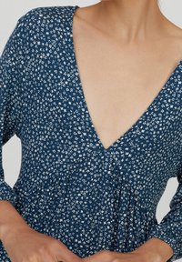 Robe fleurie bleue avec un profond décolleté en V, des manches longues et une taille froncée. Motifs de fleurs blanches contrastant avec le tissu bleu.