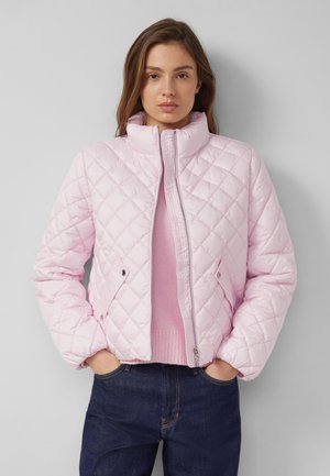 Naine kannab helepunast tikk-kuldkattega jope peal kokku sobiva kampsuniga, käed taskutes ja tumedaid siniseid teksaseid ning seisab halli tausta ees.