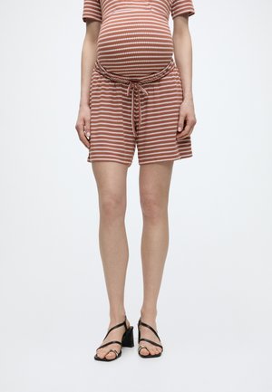 Personne enceinte portant un short et un haut en maille rayés rouille et blanc, associés à des sandales noires à talons et à brides, debout sur fond blanc.