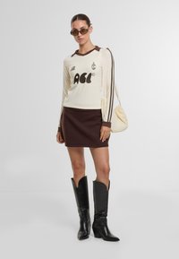 Kermainen pitkähihaista paitaa ruskeilla tehosteilla ja graafisella printillä, yhdistettynä ruskeaan minihameeseen ja mustiin polvipitkiin saappaisiin. Pieni kermainen käsilaukku.