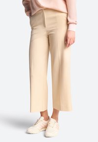 Pantalons larges beige à la texture lisse, dotés de plis à l'avant. Associés à des chaussures de sport de couleur claire avec des accents roses.