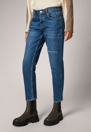 Femme portant un jean slim taille haute bleu légèrement usé et des bottines marron à semelle épaisse, debout avec les mains détendues le long du corps.