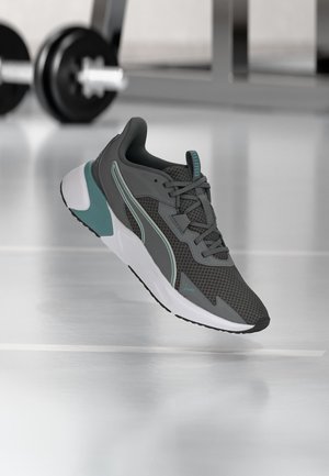 Gray and teal athletic shoe with mesh upper and white sole, set against a gym floor with blurred weights in the background.