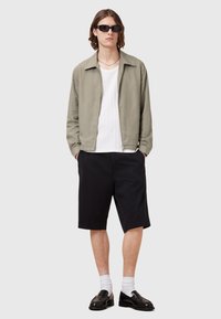 Lys olivengrøn zip-up jakke, hvid ribbet skjorte, sorte shorts, sorte loafers og hvide sokker, båret af en person med bølget hår.