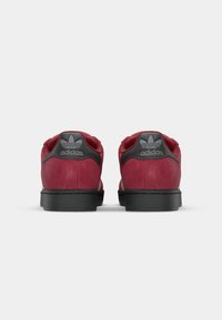 Red suede sneakers with black accents, featuring a padded collar, textured heel tab, and textured rubber sole. Classic Adidas logo visible.