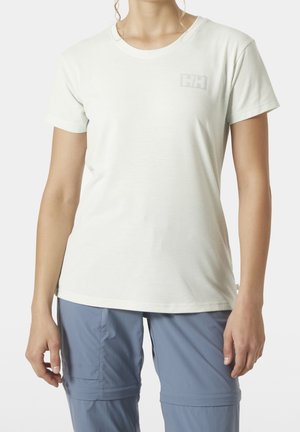 Mujer que lleva una camiseta blanca de manga corta con un logo discreto y pantalones azul claro con cremallera contra un fondo liso.