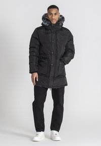 Sort pufferjakke med hætte i kunstpels, trykknaplukninger og to sidelommer. Bæres over sorte bukser og hvide sneakers.