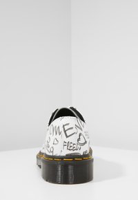 Bílé kožené boty s černými ručně psanými textovými vzory. Černá gumová podrážka s žlutým šitím. Kulatá špička a nízký profil.