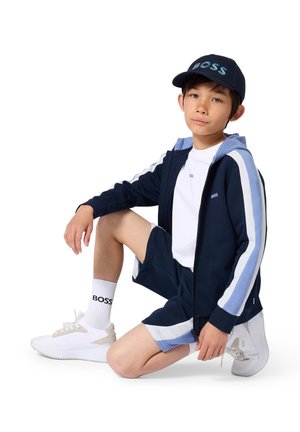 Garçon portant un survêtement bleu marine, blanc et bleu clair, des baskets blanches, des chaussettes blanches et une casquette bleu marine avec le logo "BOSS", à genoux sur un genou.