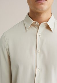 Svetlo béžová košeľa na gombíky s klasickým golierom, tromi prednými gombíkmi a hladkou textúrou; výstrih má jemný lesk.
