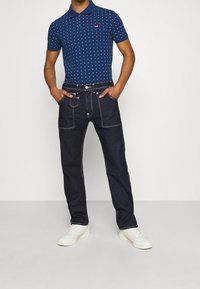 Tummansininen polo-paita, jossa on valkoinen pistekuvio ja lyhyet hihat, yhdistettynä tummiin farkkuihin, joissa on kontrastiväriset ompeleet ja sivutaskut. Valkoiset urheilukengät.