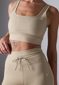 Beige treningsbh med firkantet utringning og justerbare stropper, kombinert med høyt midjeleggings som har snøring og stikkingdetaljer.