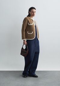 Bruine textuurjas met witte afwerkingen en zakken, gecombineerd met marineblauwe wijde pantalons. Een kleine bordeauxrode handtas maakt de outfit compleet.