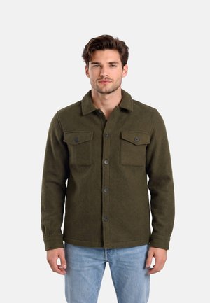 Giovane uomo che indossa una giacca verde oliva con bottoni e tasche sul petto e jeans azzurri, in piedi davanti a uno sfondo semplice.