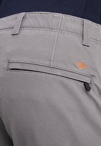 I pantaloni in tessuto grigio presentano una tasca posteriore con chiusura a zip e un logo ricamato arancione sopra la tasca. Texture liscia, vestibilità sartoriale.