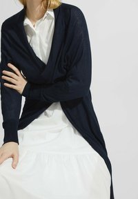 Marineblau gestrickte Strickjacke mit lässigem Schnitt, V-Ausschnitt und langen Ärmeln, kombiniert mit einem weißen Hemd und einem fließenden weißen Rock.