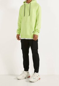 Neon vihreä huppari, jossa on kengurupocket, mustat narut, yhdistettynä mustiin cargo-housuihin ja valkoisiin urheilukenkiin.