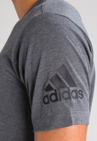 Šedé tričko s krátkým rukávem vyrobené z měkkého materiálu. Na levém rukávu se nachází černé logo Adidas. Jednoduchý, klasický design s kulatým výstřihem.