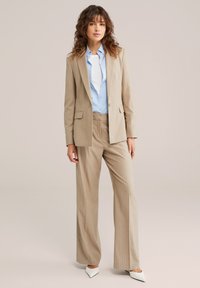 Smėlio spalvos dryžuotas atviras švarkas ir derančios kelnės, dėvimos kartu su šviesiai mėlyna marškinėlių su sagutėmis, užbaigtos baltais aštriais aukštakulniais.